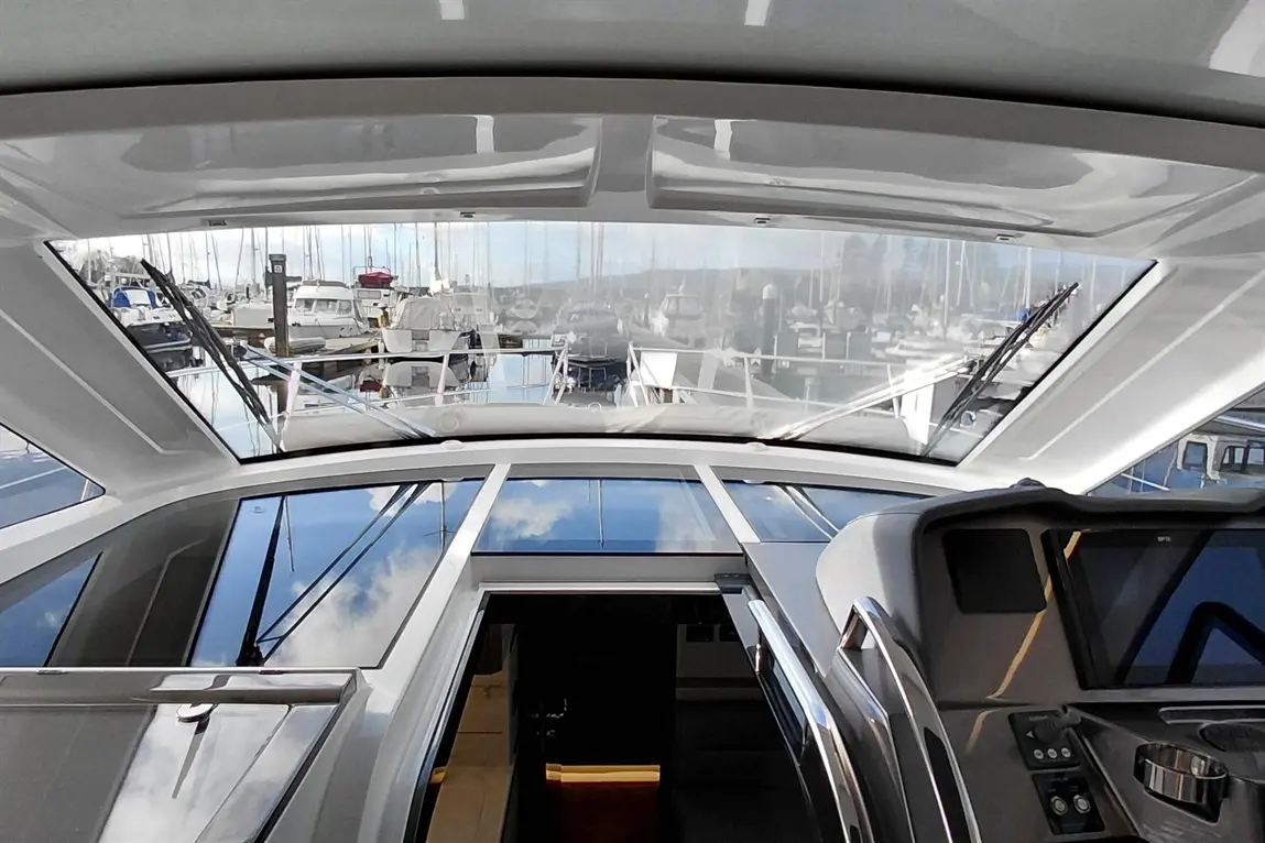 View from a yacht's helm showing boats in a marina through a glass roof, reflecting the sky and water below.